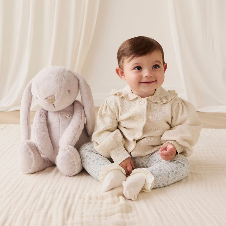 Baby sitting beside a soft beige Snuggle Bunnies plush bunny on a cream blanket, wearing a knitted cardigan and floral leggings.