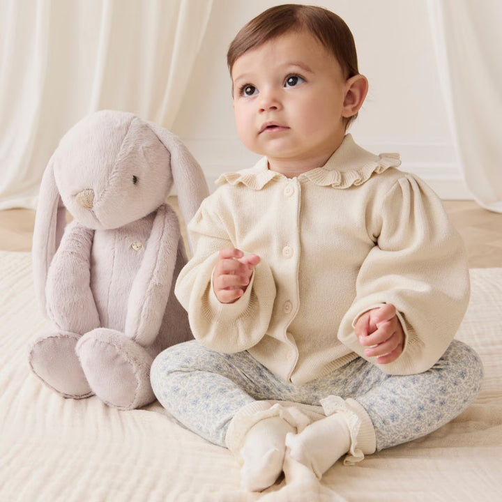 Baby sitting beside a beige Snuggle Bunnies plush bunny on a soft cream blanket.