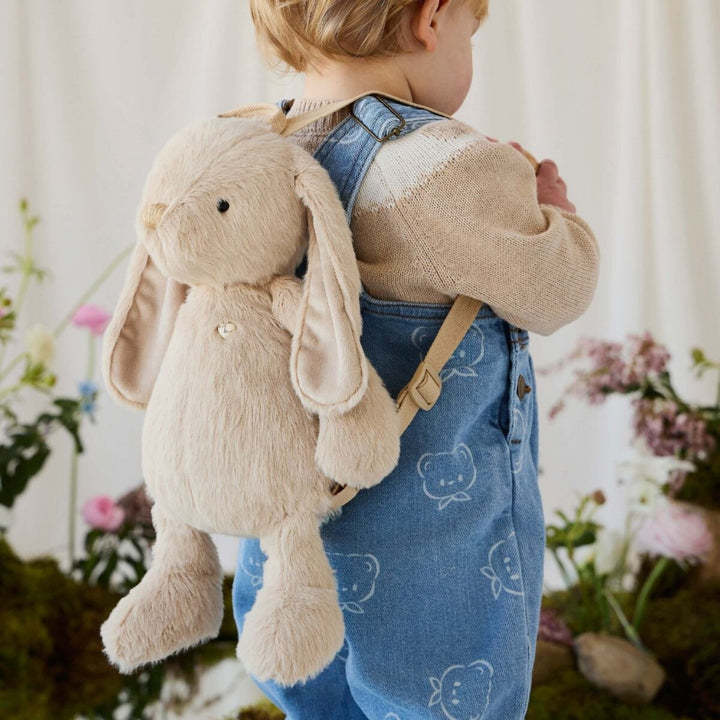 Young child wearing a soft beige bunny backpack with long floppy ears, adjustable shoulder straps and plush dangling legs, dressed in blue denim overalls.