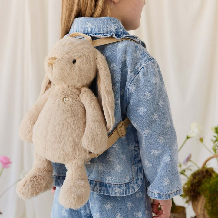 Young child wearing a hazelnut beige plush bunny backpack over a light blue floral denim outfit, showing its soft texture and adjustable straps.