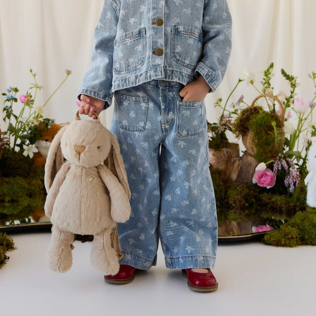 Toddler in a blue floral denim outfit and red shoes holding a soft hazelnut beige bunny backpack by its top handle, surrounded by pastel flowers.