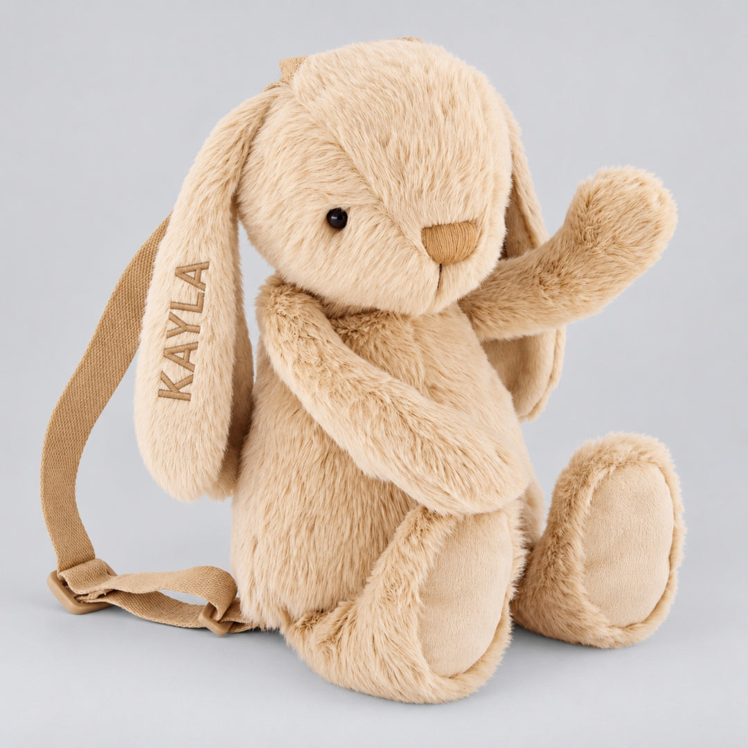 Soft hazelnut beige plush bunny backpack with floppy ears, one raised paw and the name KAYLA embroidered in beige block lettering on its ear.