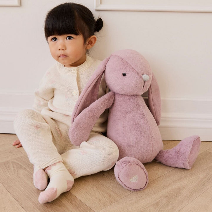 Young girl sitting beside a pink Snuggle Bunnies plush bunny on a wooden floor.