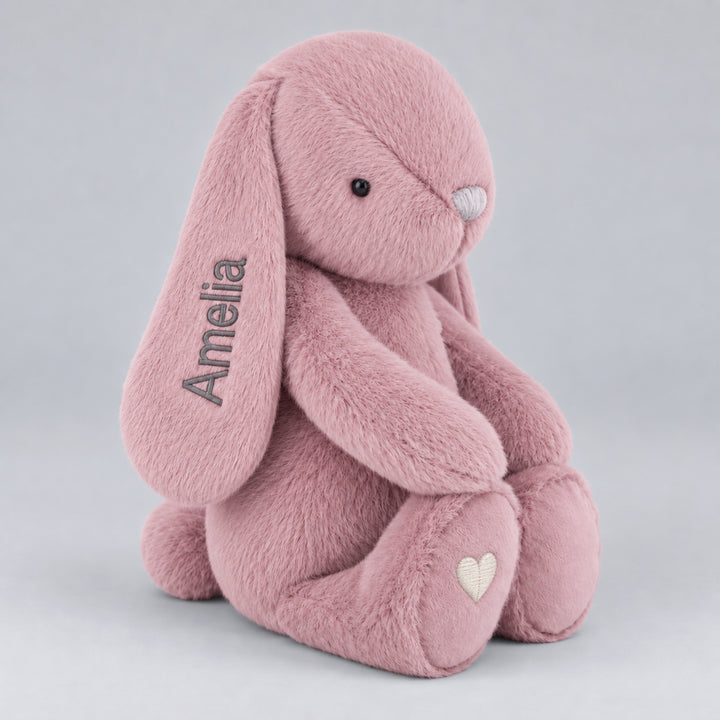 Pink Snuggle Bunnies plush bunny in side view with personalised "Amelia" ear embroidery and a cream heart on its foot.
