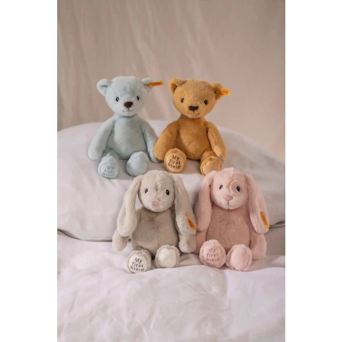 Four plush stuffed toys arranged on white bedding: two teddy bears in light blue and tan, and two bunnies in beige and pale pink. Each toy has a small yellow tag on the ear and embroidered text on the paw.