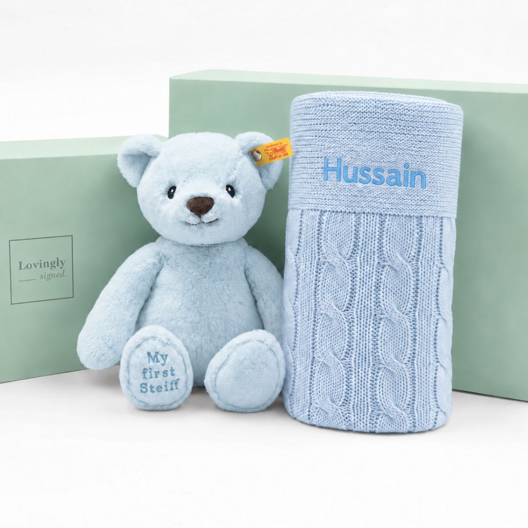 Personalised blue My First Steiff teddy bear with blue cable knit blanket embroidered “Hussain,” displayed with Lovingly Signed gift boxes.
