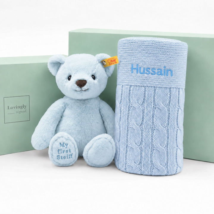 Personalised blue My First Steiff teddy bear with blue cable knit blanket embroidered “Hussain,” displayed with Lovingly Signed gift boxes.