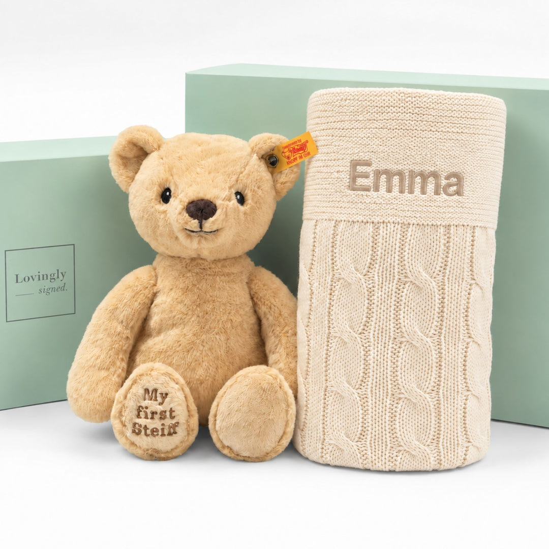 Personalised My First Steiff teddy bear with cream cable knit blanket embroidered “Emma,” displayed with Lovingly Signed gift boxes.