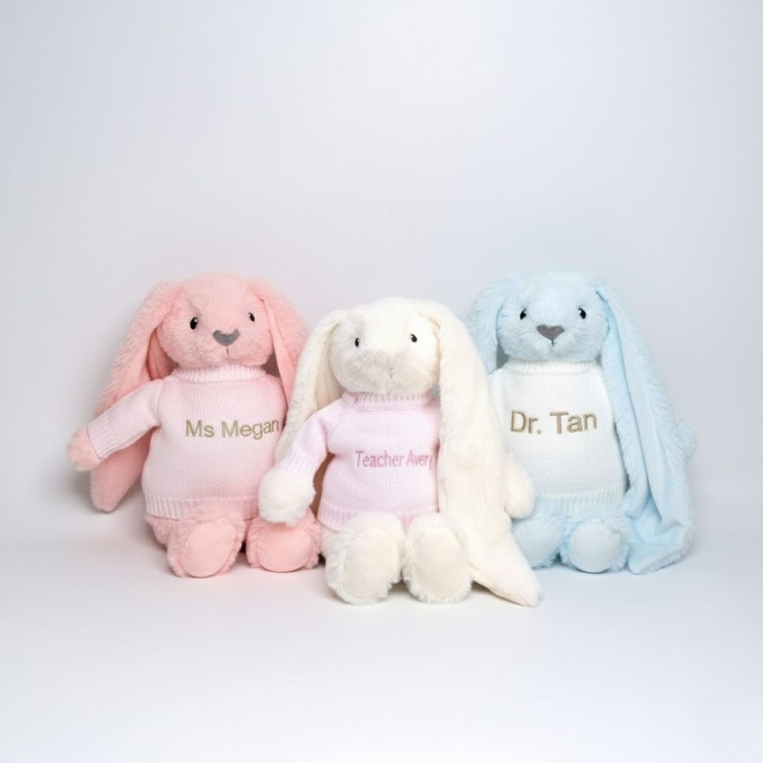 Three plush bunnies in pink, white, and blue with embroidered names on a light gray background