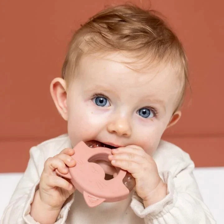 A baby biting and holding a dusty pink silicone teether against their teeth, wearing a cream-colored knit top against a solid terracotta background.
Trixie Mrs Cat Cooling Baby Teether